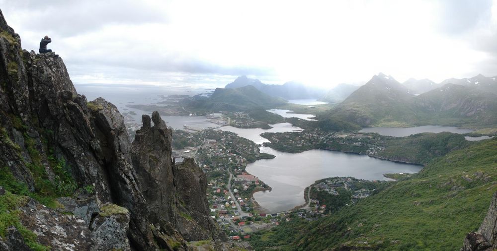 A panorama from high up shows on the left the rocky formation called the Svolvær Goat. It sits in front of a landscape with a town, Svolvaer, which is situated among bays, hills, and mountains.