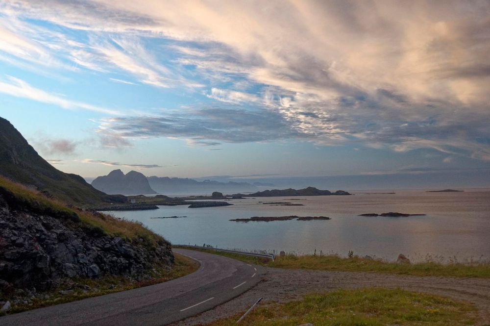 An empty road carves around the edge of a mountain on the left. The view is that of the calm sea with several islands in it. The sky has a cloud in it, and the light is very low.
