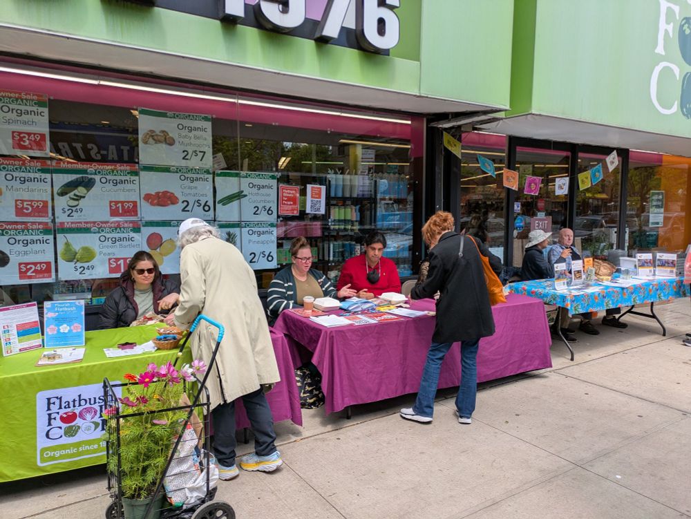 In front of a grocery store are three tables, each of which has flyers and people talking to passersby. The middle table has Flatbush Streets for People.