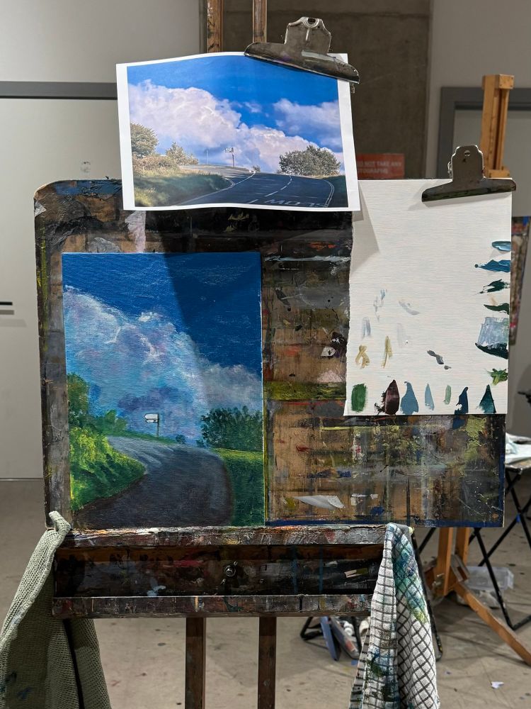 Picture of an Acrylic painting on an easel in a studio. The painting is of a road junction with big clouds across the bottom of the horizon. A sign post points off to the left