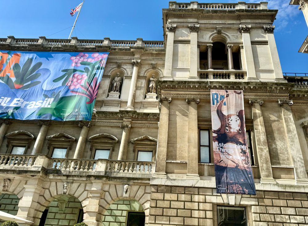 Outside of the Royal Academy in London on a sunny blue sky day with banners advertising their exhibitions. The Victor Hugo one has an ink drawing of an Octopus