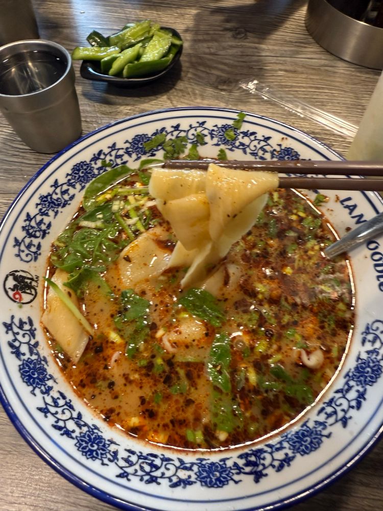 bowl of soup with vegetables and chopsticks picking up a pair of large flat noodles