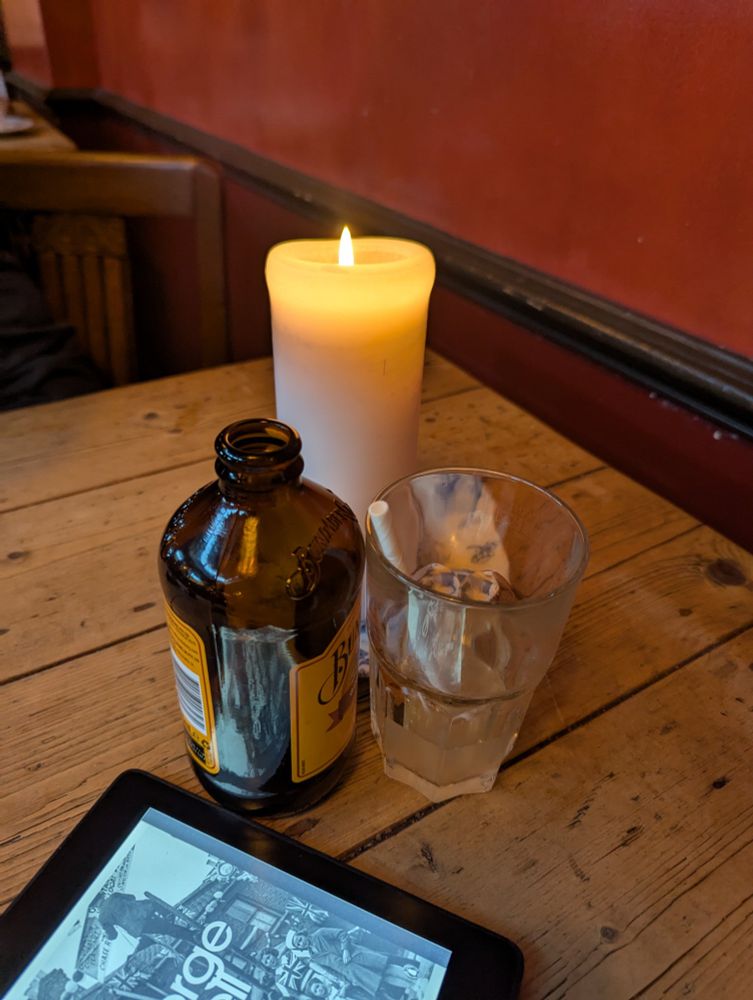 A scrubbed wooden table with an empty ginger beer bottle, a near empty glass with a paper straw, a candle and the corner of my Kindle. 
