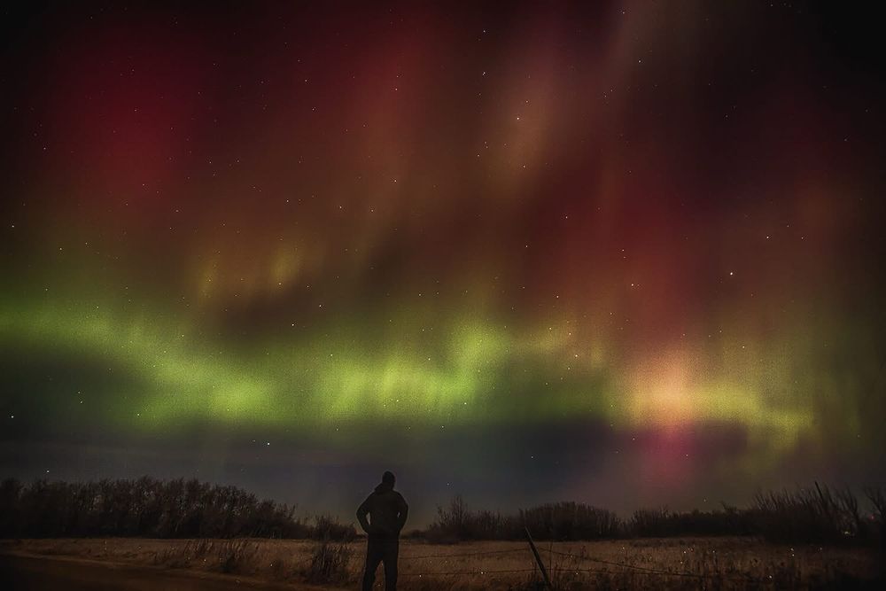 Silhouette of a man standing in the middle of the frame. Night time, northern lights dancing in the sky. 
