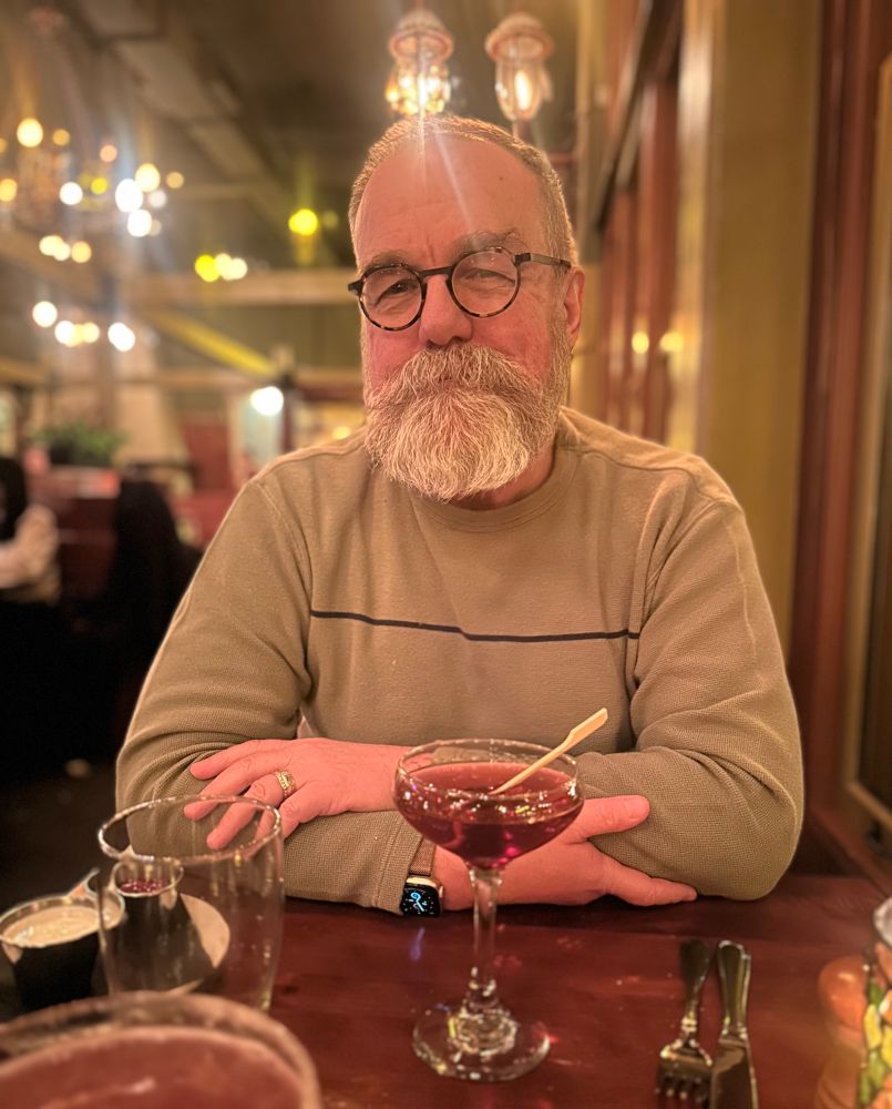 Man with round glasses and a gray beard with arms crossed in front of a Manhattan. It’s his birthday.