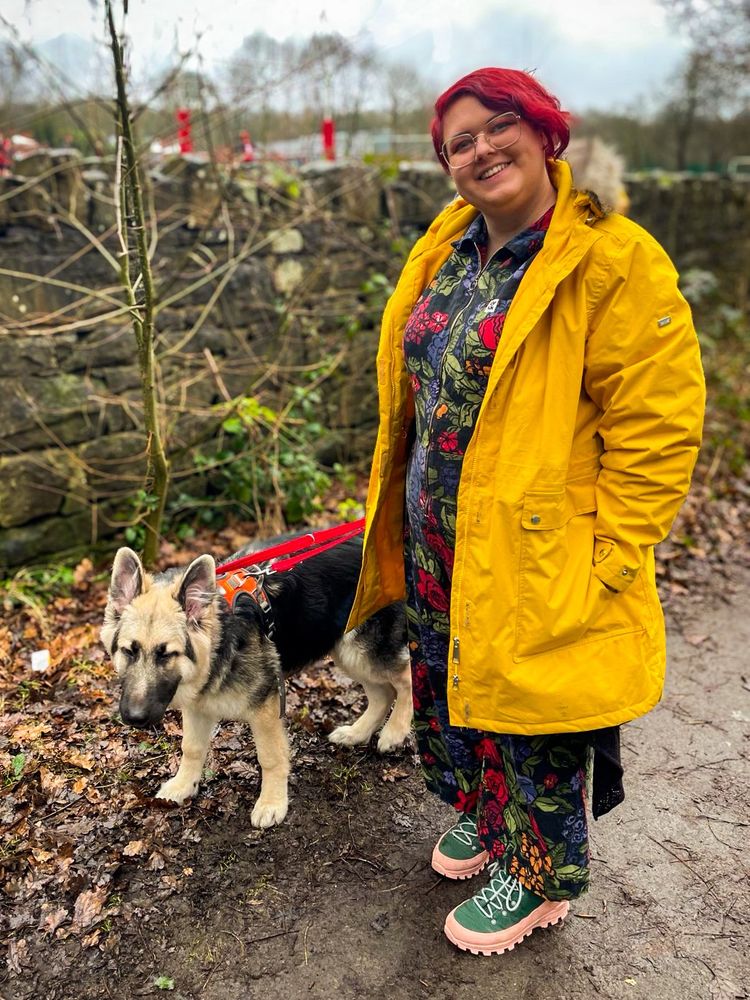 A woman with short red hair wearing a yellow outdoor coat stands smiling next to a very large German Shepherd puppy