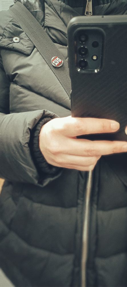 Fahrstuhl-Selfie mit schwarzer Winterjacke und FCK AFD Pin am Taschengurt.