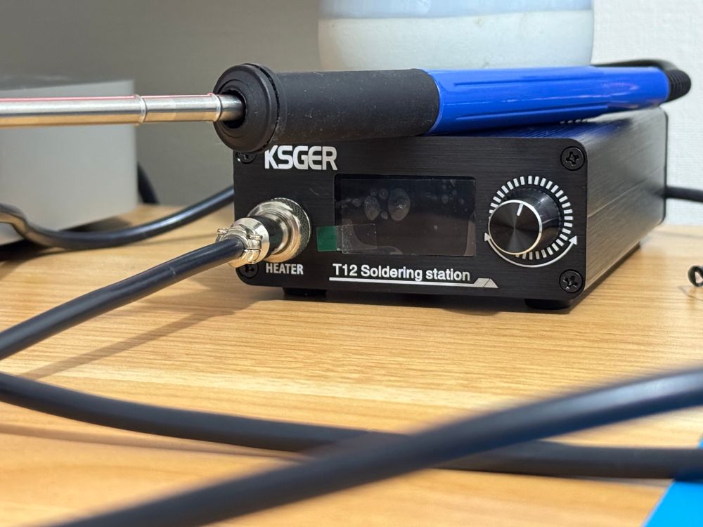 Un boitier avec une sortie estampillée HEATER, un écran sur le boitier avec en dessous d’écrit “T12 soldering station”. Un bouton qui tourne à droite.
Repose sur le boitier un fer à souder avec un manche bleu.
Le fil court sur toute la table en bois clair.