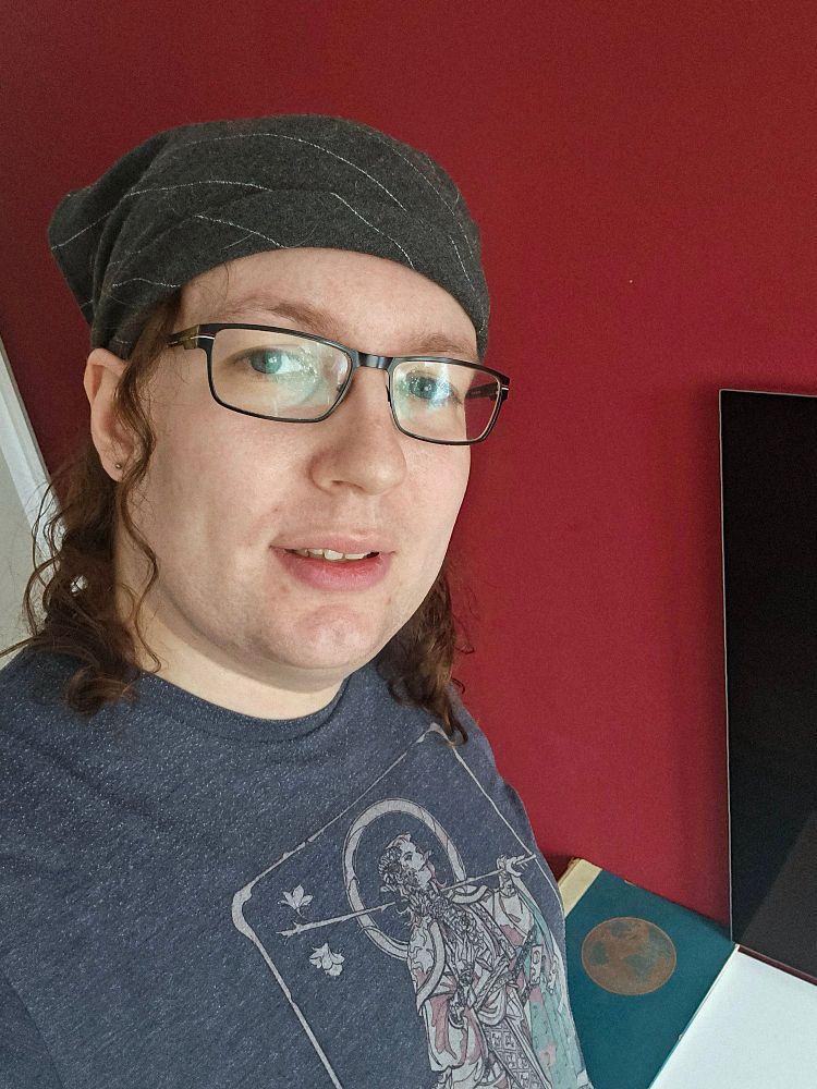 WHOA IT'S ALICE!
selfie of a white femme with glasses and light brown curly hair, looking invitingly at the camera. she wears a black headscarf and a gray-blue shirt with a character from critical role S2 on it (don't ask me who, i forgot). she stands in front of a red painted wall. barely visible under the shirt are what she thinks of as her great set of tits.