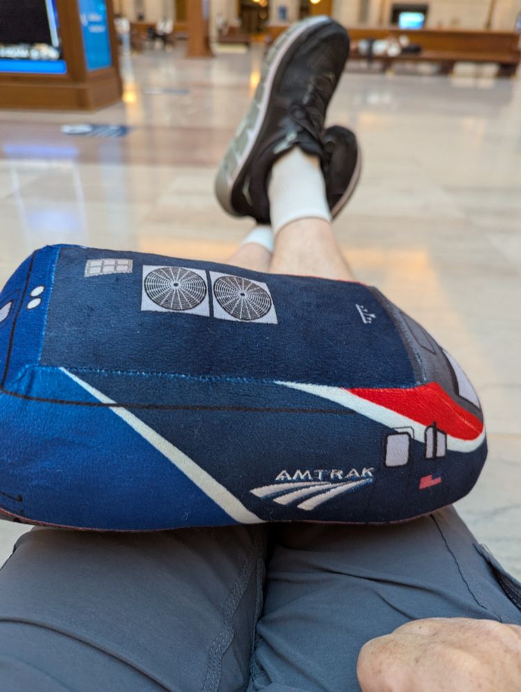 Stuffed dark blue Amtrak locomotive with a thick red stripe across the nose sitting on my lap as I have my feet propped on my suitcase.