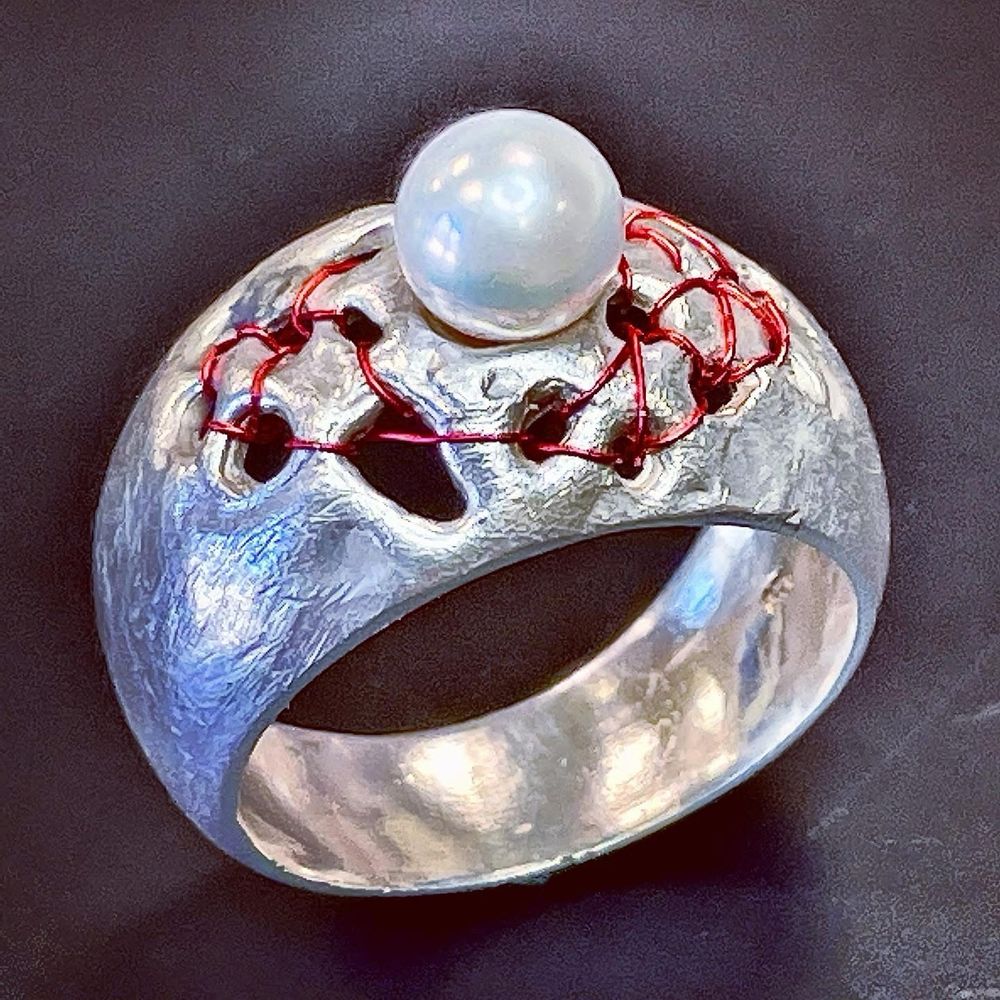 A unique silver ring features a prominent pearl at the top, embellished with intricate red thread woven through a textured design. The ring has an artistic, modern look with contrasting colors and textures.