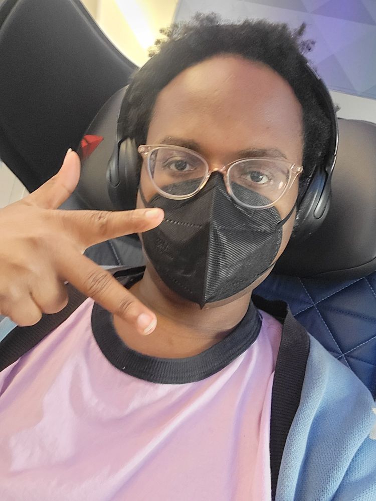 A cute black trans girl wearing a face mask while in first class on her flight. Is smiling but you can't see it, lol. Holding up two fingers and her thumb (I don't know what that's supposed to mean!)