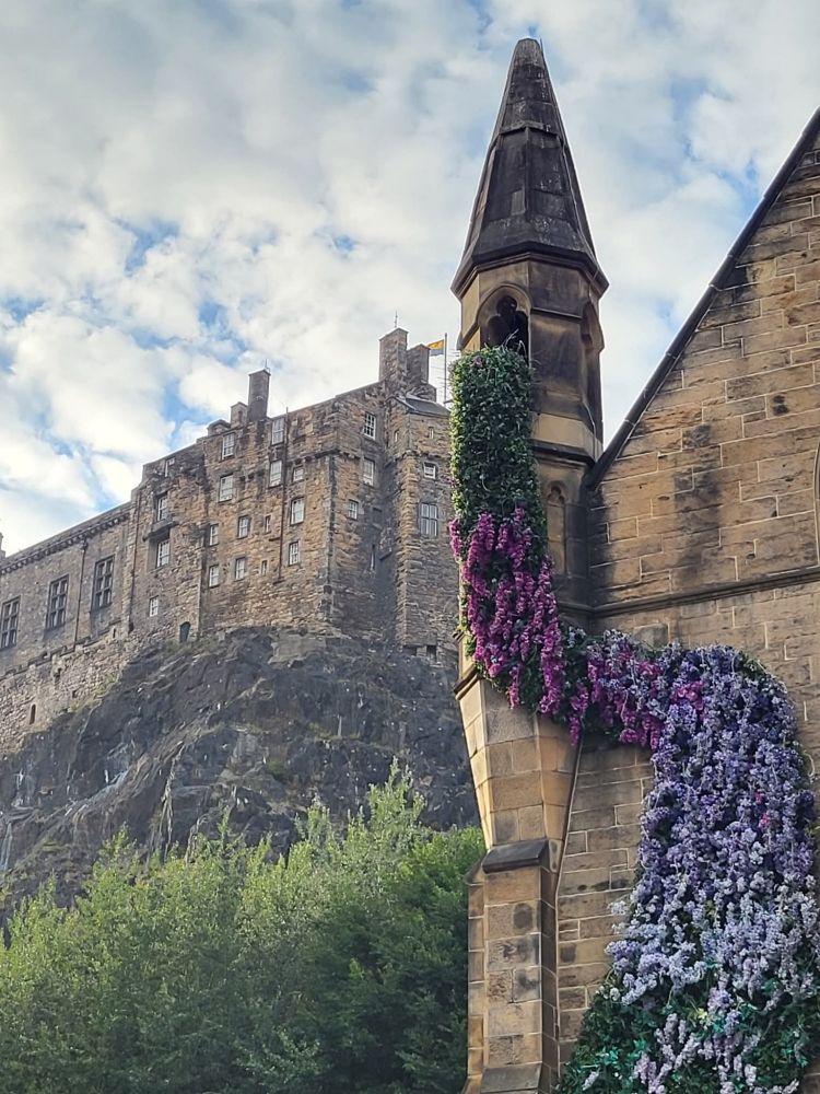 Looking up at Edinburgh Castle from a street below. The building in the foreground looks like it has a tower with wisteria flowing out of it and down the building. At the top of the tower the flowers are a deep purple, but the colour fades to a light lilac colour as they go closer to the ground. It's beautiful 