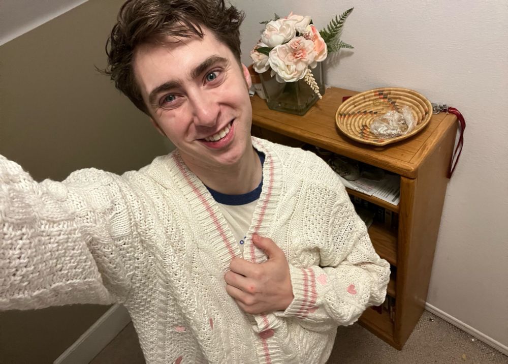 Will is smiling while holding the chest area of his cardigan with one hand. The cardigan that is a creamy white cardigan with light pink hearts stitched around it with a pink line around the neck and button holes with pink hearts as buttons