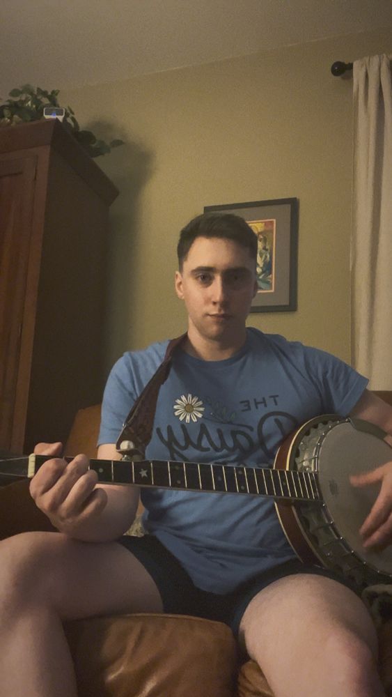 Will in a blue shirt and black shorts holding a banjo that once belonged to his grandpa and is sitting on a brown leather couch
