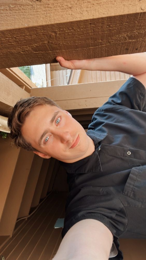 Will in a black button up short sleeve shirt with one arm on a wooden post and standing  under a wooden shed 