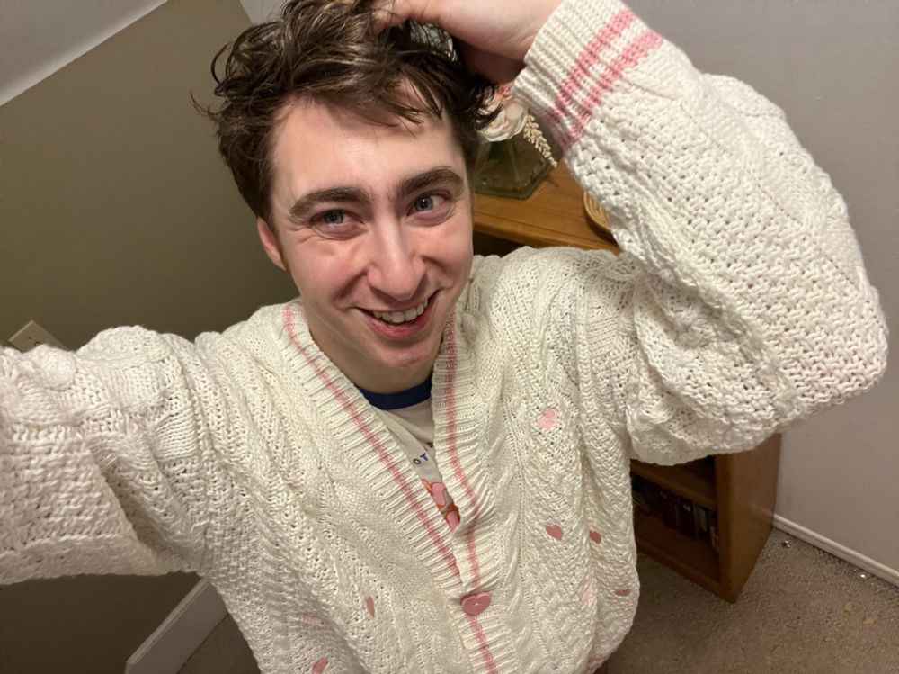 Will smiling wearing his Lover cardigan with one hand touching his hair. The cardigan that is a creamy white cardigan with light pink hearts stitched around it with a pink line around the neck and button holes with pink hearts as buttons