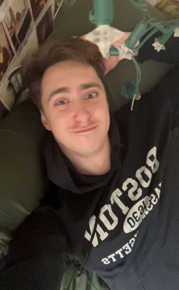 Will laying on his bed wearing a black hoodie with Boston Massachusetts on it in white letters with one hand behind his head. His bed sheets and pillow are a forest green color 