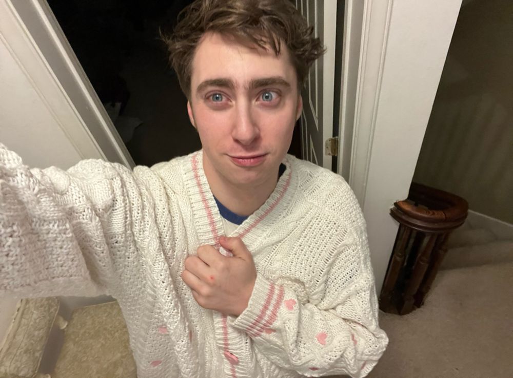 Will with a closed mouth smile holding the chest area of his Lover cardigan. The cardigan that is a creamy white cardigan with light pink hearts stitched around it with a pink line around the neck and button holes with pink hearts as buttons