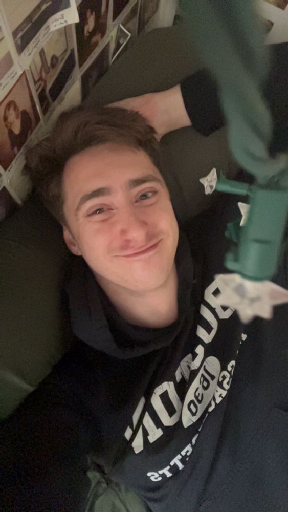 Will laying on his bed with a smirk on his face and one arm behind his head, he is wearing a black hoodie with Boston Massachusetts on it in white letters 