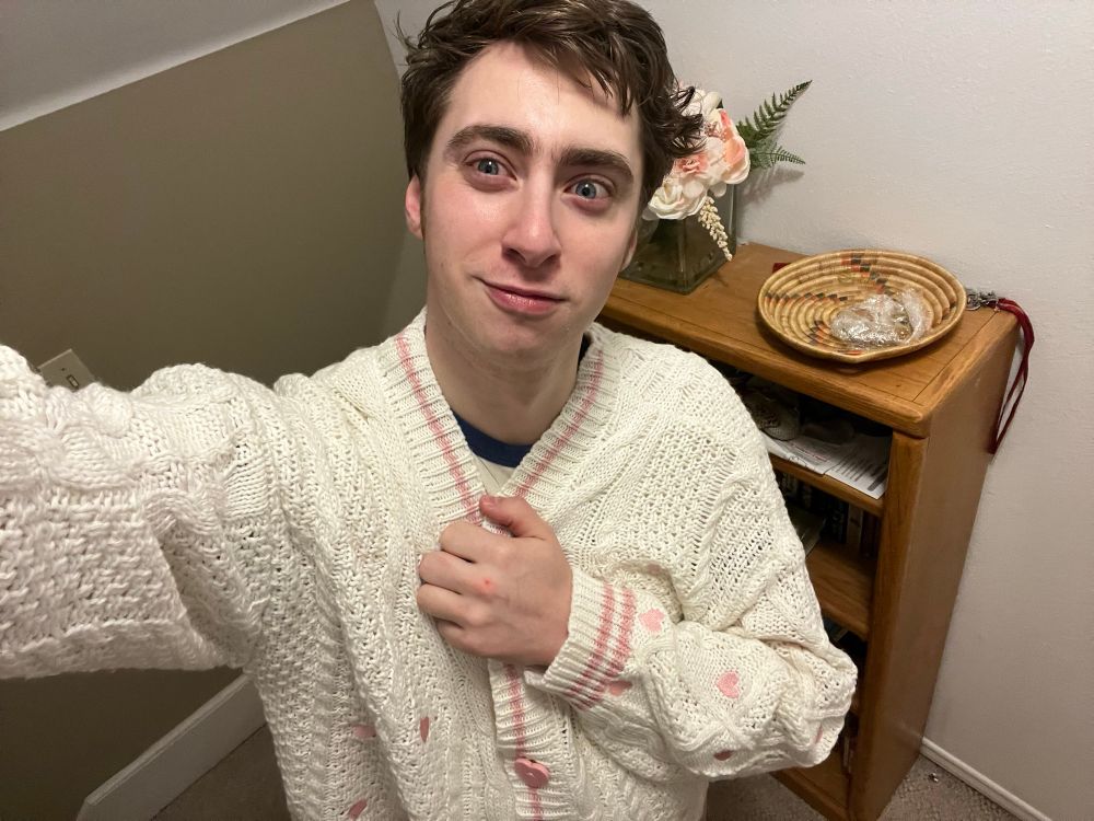 Will with a closed mouth smile wearing his Lover cardigan! The cardigan that is a creamy white cardigan with light pink hearts stitched around it with a pink line around the neck and button holes with pink hearts as buttons