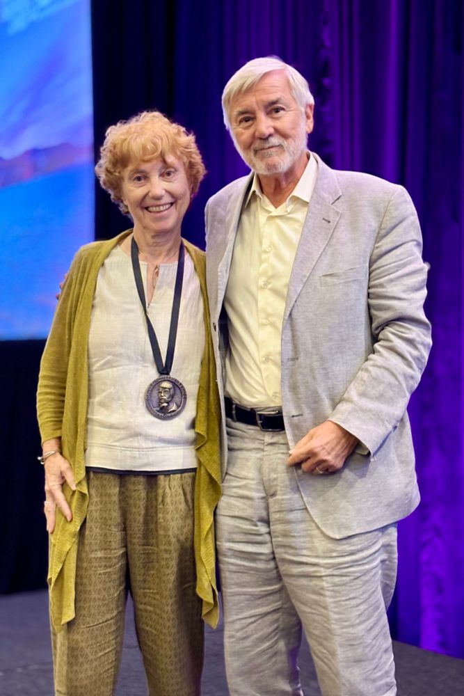 Nora S. Newcombe and Robert Glushko stand together smiling on stage at CogSci 2025. Nora wears the Rumelhart Prize medal around her neck