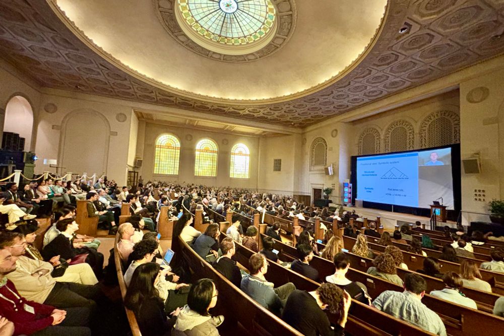A large audience fills a historic hall with wooden pew seating, ornate ceiling details, and a stained-glass dome. People face a stage with a large screen displaying a presentation, as the Rumelhart 25th Anniversary Event takes place at the Internet Archive