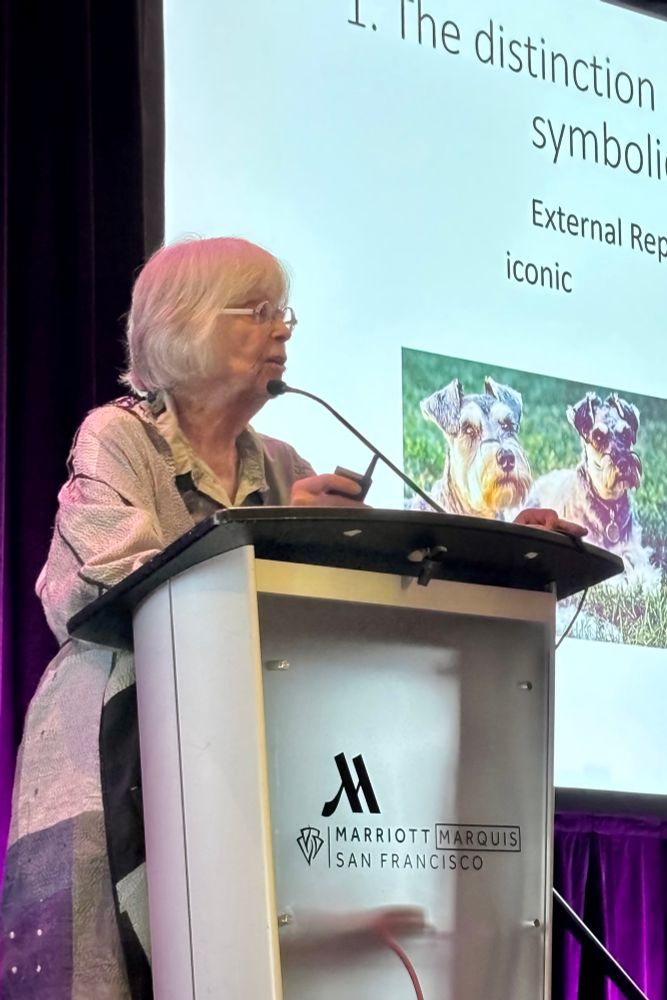 Susan Carey speaks at a lectern with the Marriott Marquis San Francisco logo during her keynote at CogSci 2025. Behind her, a presentation slide shows  an image of two dogs in a grassy field.