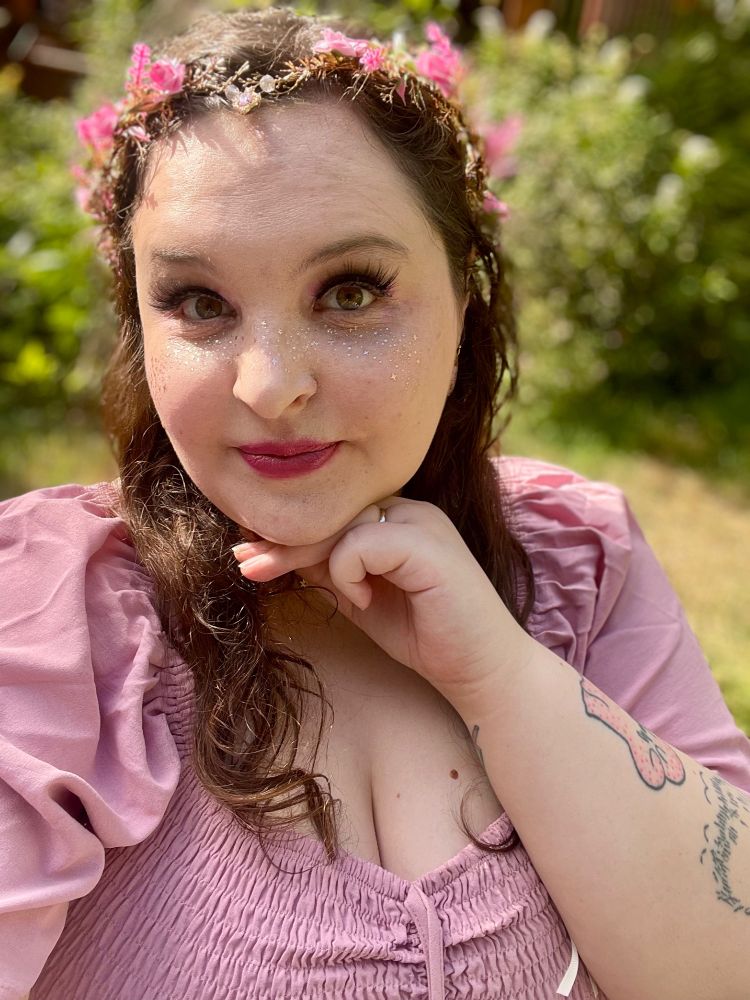 Selfie of me (white femme with brown hair) in a pink dress, a pink flower crown, and makeup in gold and pink tones.