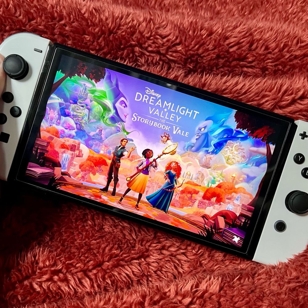 Image is of a white Nintendo switch with the new storybook vale expansion pack on the Disney Dreamlight Valley game on the display. Behind, is a rust coloured fluffy throw.