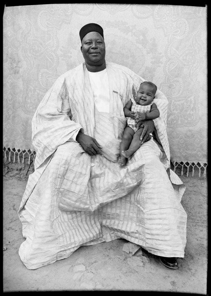 a baby with very round cheeks sitting on her father's lap in a black and white portrait by Seydou Keita. Her father is wearing a white grand boubou and a black hat; she's wearing a checkered onesie. They are sitting on a stool in front of an elaborately embroidered cloth. They both look absolutely delighted.