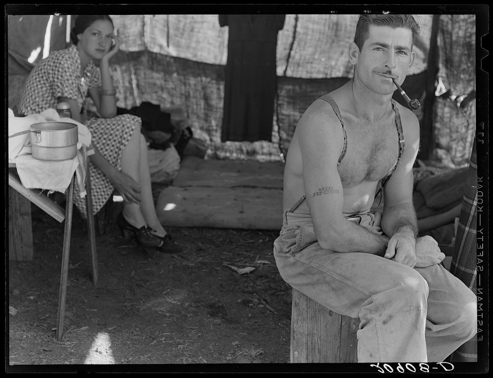 Black and white photo shows a seated, shirtless, mustachioed man wearing suspenders and jeans, smoking a pipe, flexing his arm with his Social Security number tattooed on it. His wife is in the background, cupping her head in her hand.

Unemployed lumber worker goes with his wife to the bean harvest. Oregon. Photographed by Dorothea Lange, U.S. Farm Security Administration/Office of War Information Black & White Photographs. Library of Congress Prints and Photographs Division.