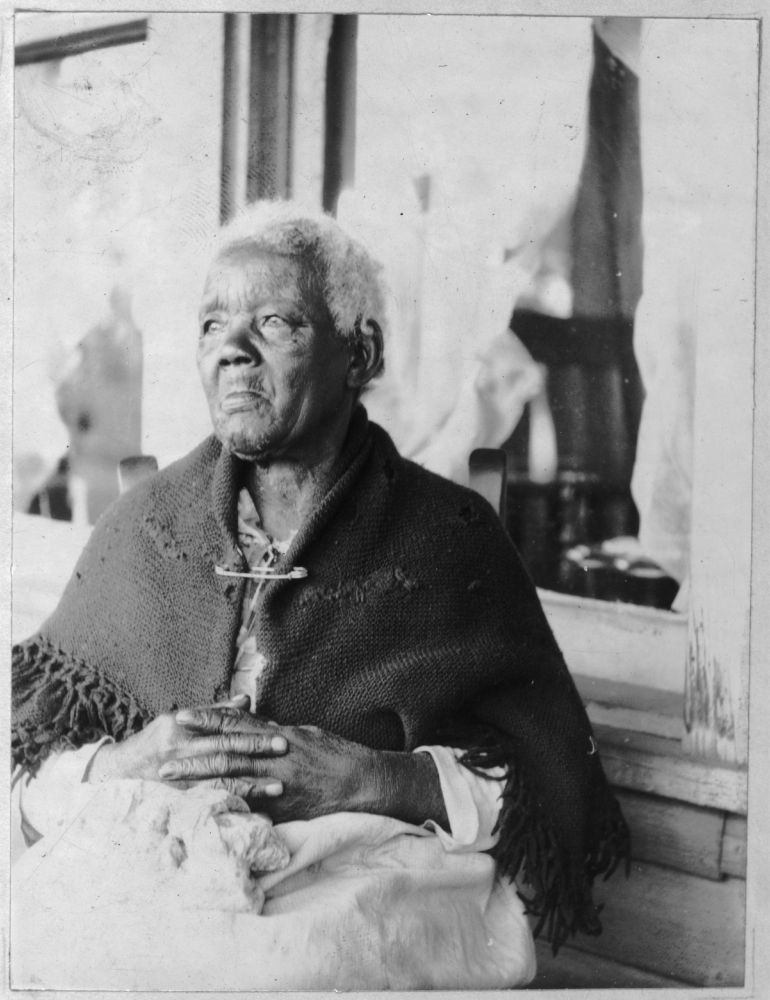 A portrait of an elderly Black woman, Sarah Gudger, who was reportedly 121 years old at the time the photo was taken to accompany the narrative she shared with the Federal Writers' Project. Her hair is short and white, her hands are in her lap with her fingers laced together and she is looking into the distance, to the left of the camera (her right). Her tattered shawl is safety-pinned together under her neck.

