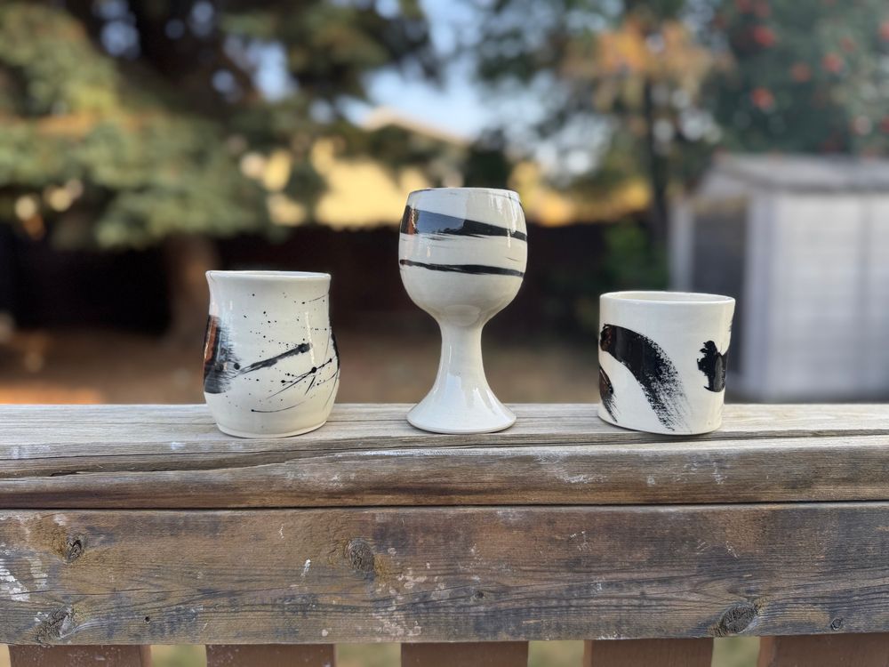 From left to right three cups on an exterior fence: the first a splattered black and white curvy cup, the second a chalice with organic stripes of black and white, and a straight wall cup with organic streaks of black and white.