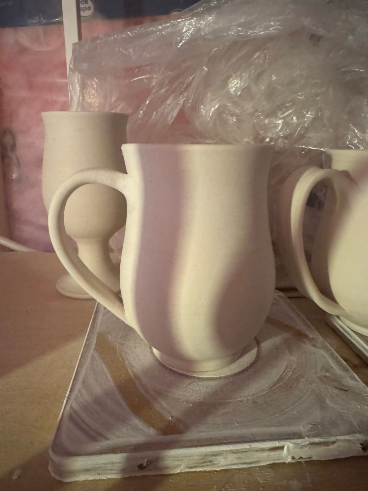 A closeup view of a tall and curvy porcelain mug. In the background other mugs, a chalice and pink insulation are visible.