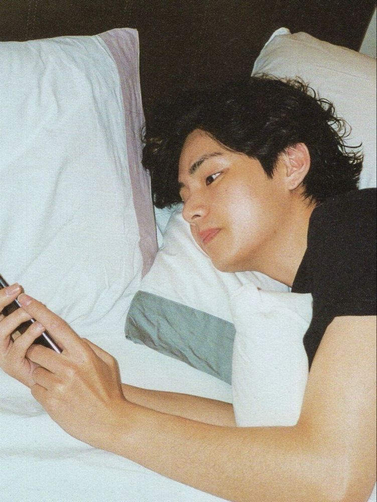 taehyung lying in bed on his stomach, looking at his phone while cuddling a pillow