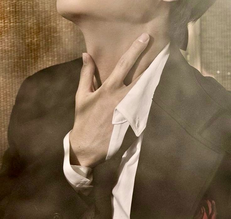 A close up photo of Kim Taehyung wearing a black suit. He’s loosely grabbing the base of his throat. 