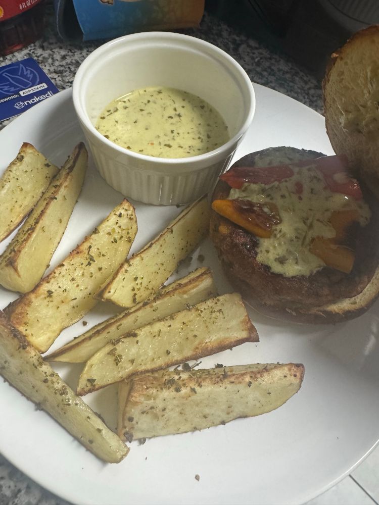 The same plate, from a different angle. There is aioli in the ramekin. The top bun of the burger is lifted off, revealing more aioli and red and yellow sweet peppers with a light char