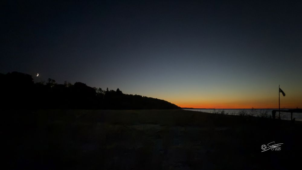 A photo of a Lake Superior sunset at Grand Marais. The horizon glows orange against the fading blue sky, with a crescent moon visible above the darkened treeline and a flag silhouetted on the right.