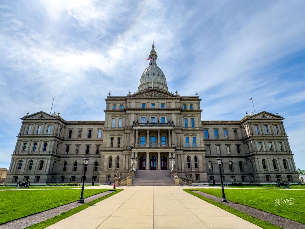 Front view of the Michigan State Capitol building in Lansing on a sunny day, showing its central dome, symmetrical neoclassical architecture, and a wide concrete walkway leading up through a green lawn.