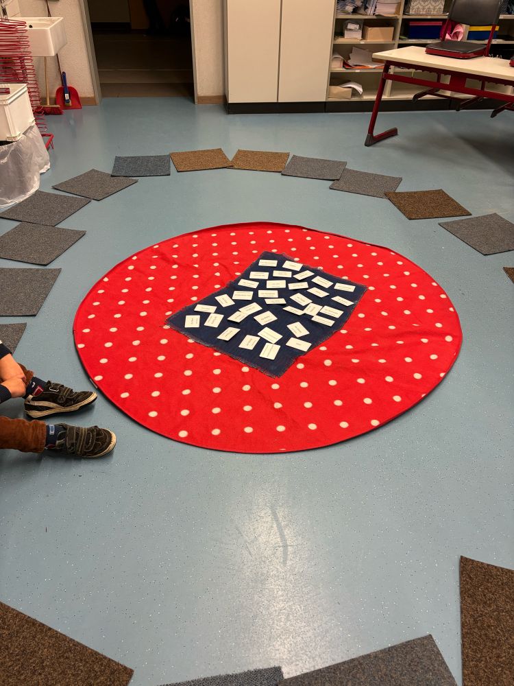 Das Foto zeigt einen hellen Raum mit einem runden, roten Teppich in der Mitte. Der Teppich hat weiße Punkte und darauf liegt ein dunkelblaues Tuch mit vielen kleinen, weißen Kärtchen, die gleichmäßig darauf verteilt sind. Um den Teppich herum sind quadratische Teppichfliesen in verschiedenen Grautönen und Brauntönen im Kreis ausgelegt. Am Rand des Bildes sind die Füße eines Kindes zu sehen, das auf dem Boden sitzt.