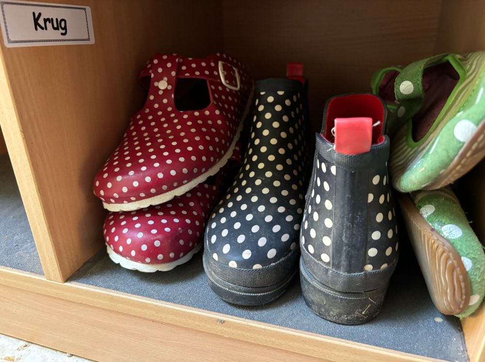 Auf dem Bild sieht man ein Holzschuhregal mit einem Fach, das mit dem Namensschild “Krug” beschriftet ist. In diesem Fach befinden sich drei Paar Schuhe: ein Paar rote Hausschuhe mit weißen Punkten, ein Paar blaue Gummistiefel mit weißen Punkten und ein weiteres Paar grüne Hausschuhe mit weißen Punkten.