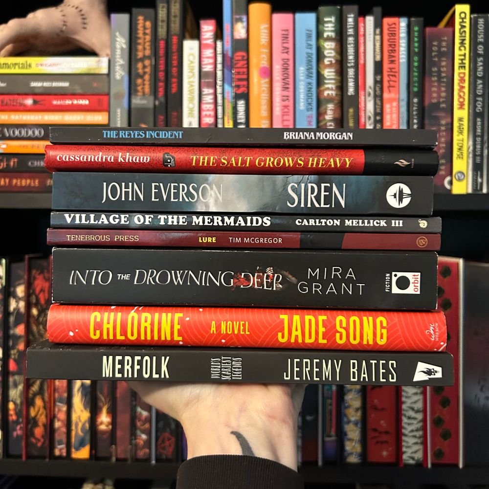 A hand holds a stack of ocean-themed and mermaid horror books in front of a colorful bookshelf. Titles include: The Reyes Incident by Briana Morgan, The Salt Grows Heavy by Cassandra Khaw, Siren by John Everson, Village of the Mermaids by Carlton Mellick III, Lure by Tim McGregor, Into the Drowning Deep by Mira Grant, Chlorine by Jade Song, and Merfolk by Jeremy Bates.