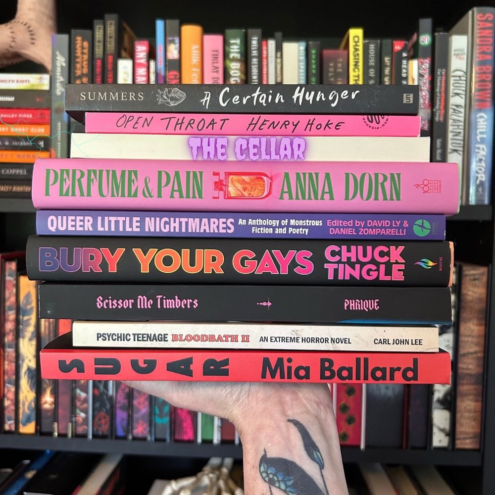 A hand with a whale tattoo holds a stack of bold, queer, and horror-tinged books in front of a dark bookshelf. Titles include A Certain Hunger, Open Throat, The Cellar, Perfume & Pain, Queer Little Nightmares, Bury Your Gays, Scissor Me Timbers, Psychic Teenage Bloodbath II, and Sugar. The spines pop in neon pinks, purples, and reds—serving queer chaos, monstrous femininity, and extreme horror realness.