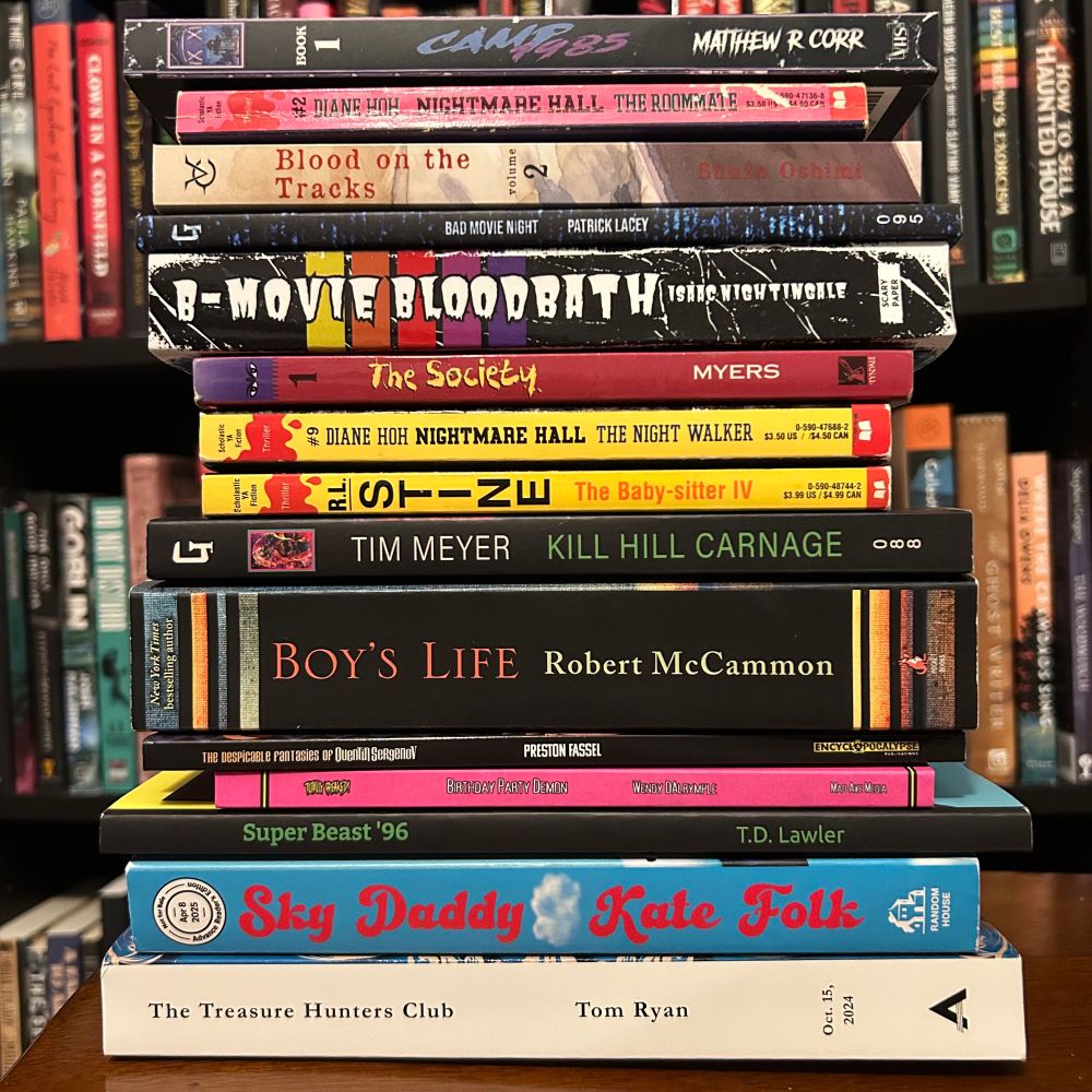 A stack of horror and thriller books arranged on a wooden table in front of a blurred bookshelf. Titles from top to bottom include: Camp 85 by Matthew R. Corr, Nightmare Hall: The Roommate by Diane Hoh, Blood on the Tracks by Shuzo Oshimi, Bad Movie Night by Patrick Lacey, B-Movie Bloodbath by Isaac Thorne, The Society by Myers, Nightmare Hall: The Night Walker by Diane Hoh, The Baby-sitter IV by R.L. Stine, Kill Hill Carnage by Tim Meyer, Boy’s Life by Robert McCammon, The Despicable Fantasies of Quentin Sergenov by Preston Fassel, Birthday Party Demon by Wendy Dalrymple, Super Beast ’96 by T.D. Lawler, Sky Daddy by Kate Folk, and The Treasure Hunters Club by Tom Ryan.