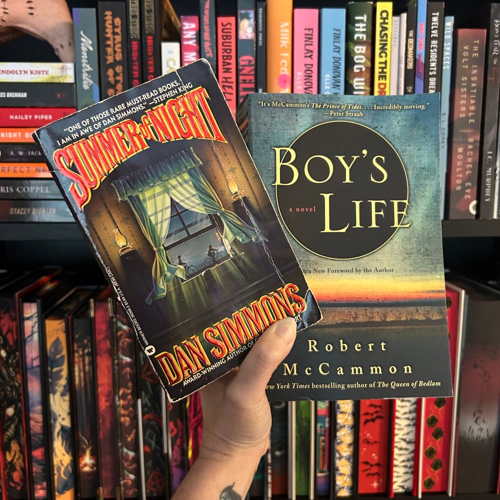 A hand holds up two books in front of a bookshelf filled with colorful spines. The left book is “Summer of Night” by Dan Simmons, featuring a vintage-style cover with a glowing window and children outside. The right book is “Boy’s Life” by Robert McCammon, with a more subdued cover showing a sunrise or sunset over water. The background includes various horror and fiction titles arranged on the shelves.