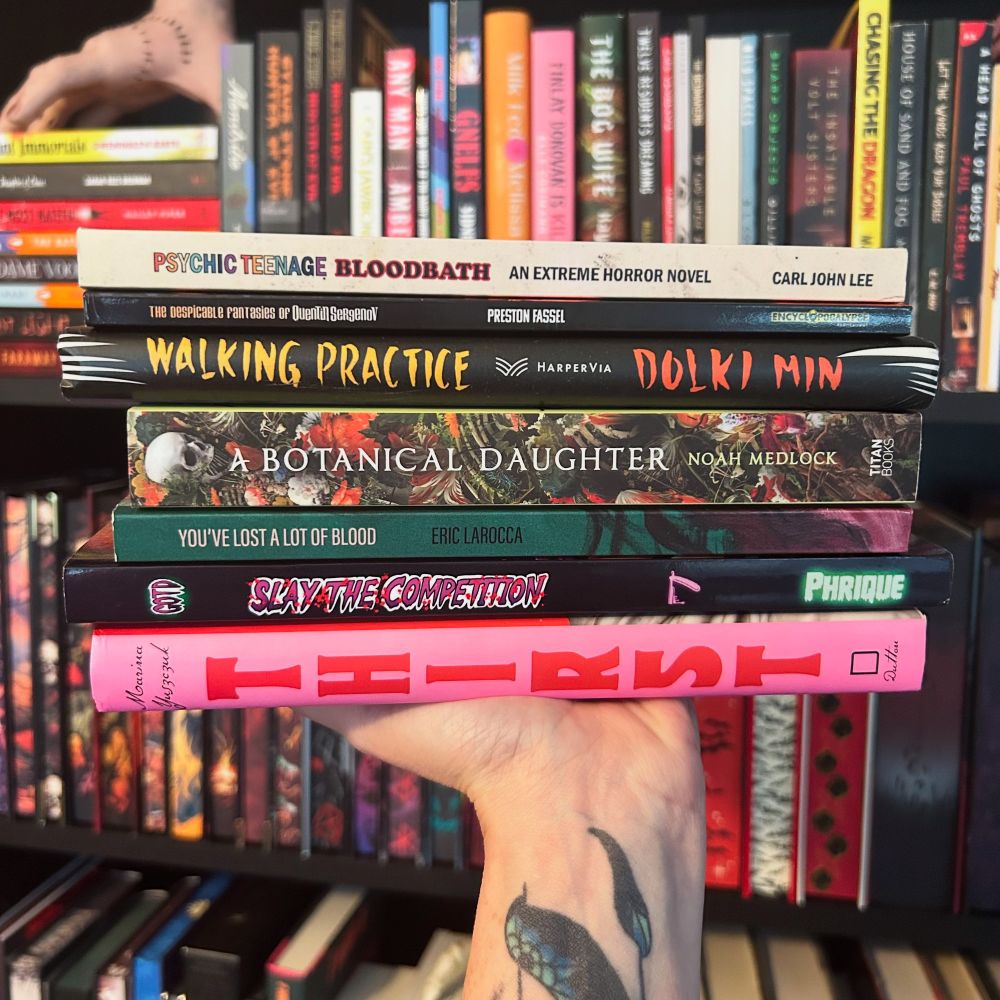 A hand with a floral tattoo holds a vibrant stack of horror and speculative fiction books in front of a colorful bookshelf. The titles from top to bottom are: “Psychic Teenage Bloodbath” by Carl John Lee, “The Despicable Fantasies of Quentin Sergenov” by Preston Fassel, “Walking Practice” by Dolki Min, “A Botanical Daughter” by Noah Medlock, “You’ve Lost a Lot of Blood” by Eric LaRocca, “Gig of the Damned: Slay the Competition!” by Phrique, and “Thirst” by Marina Yuszczuk. The books are visually striking, showcasing a blend of bold fonts, neon hues, and eerie cover art.