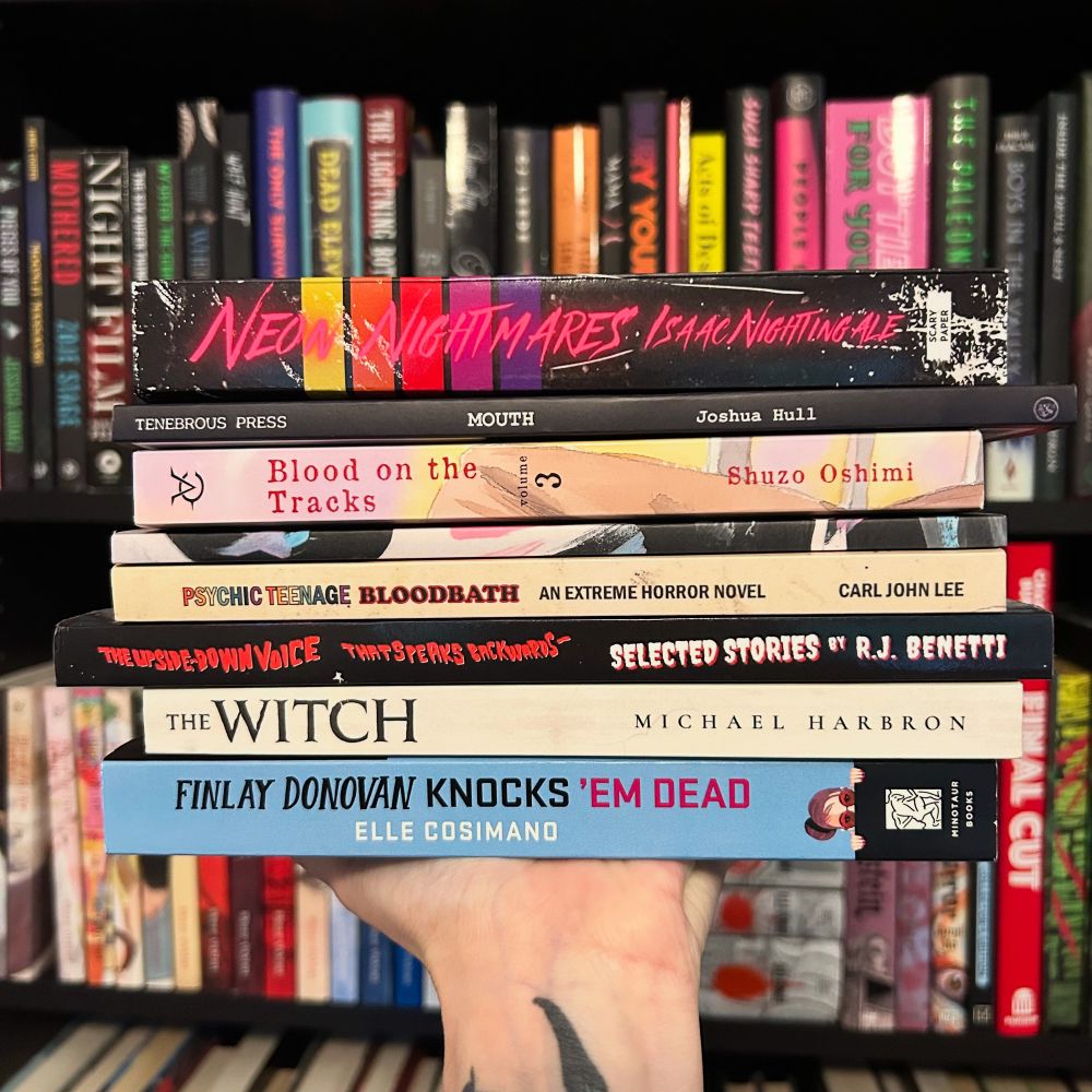 A tattooed hand lifts a stack of seven eerie and eclectic books in front of dark, moody shelves lined with horror and thriller titles. The stack includes neon-drenched nightmares, unsettling manga, extreme horror, and witchy tales: Neon Nightmares by Isaac Nightingale, Mouth by Joshua Hull, Blood on the Tracks Vol. 3 by Shuzo Oshimi, Mortifier by Sarah Derosa, Psychic Teenage Bloodbath by Carl John Lee, The Upside-Down Voice That Speaks Backwards: Selected Stories by R.J. Benetti, The Witch by Michael Harbron, and Finlay Donovan Knocks ’Em Dead by Elle Cosimano. The backdrop of spines in blacks, reds, and neons adds to the haunted library aesthetic.