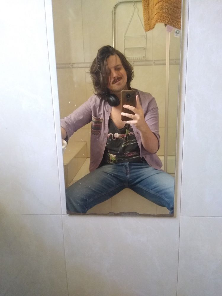 Bathroom selfie of me sitting on the edge of my bathtub. I'm wearing a lavender shirt over a black singlet with a pattern of flamingos. Also I am wearing jeans