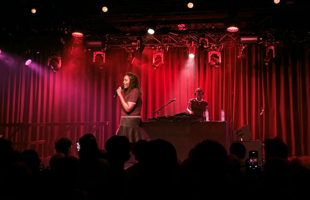 noname, smiling and holding the mic, on stage with red lights